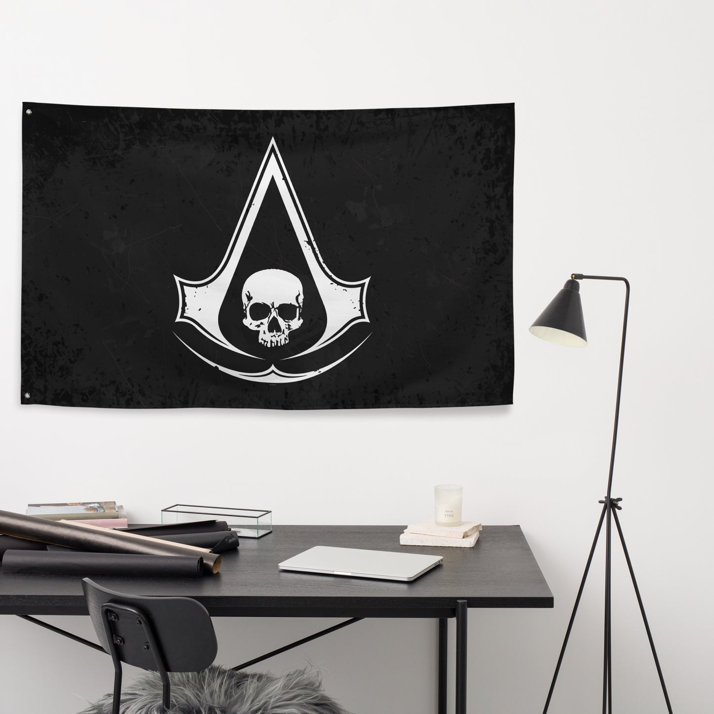 A black Assassin's Creed flag hangs above a dark desk with a laptop, candle, and rolls of paper, next to a black floor lamp.