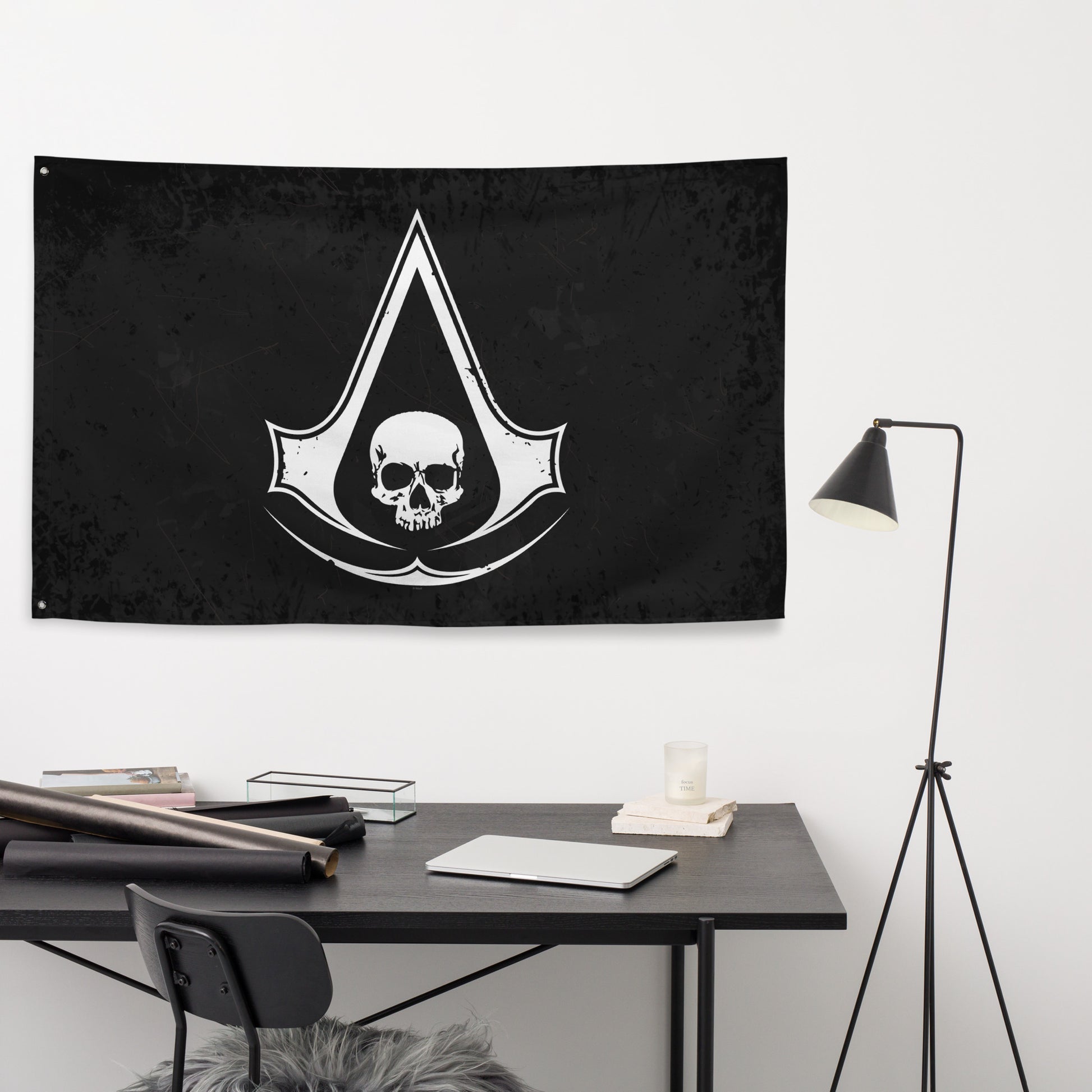 A black Assassin's Creed flag hangs above a dark desk with a laptop, candle, and rolls of paper, next to a black floor lamp.