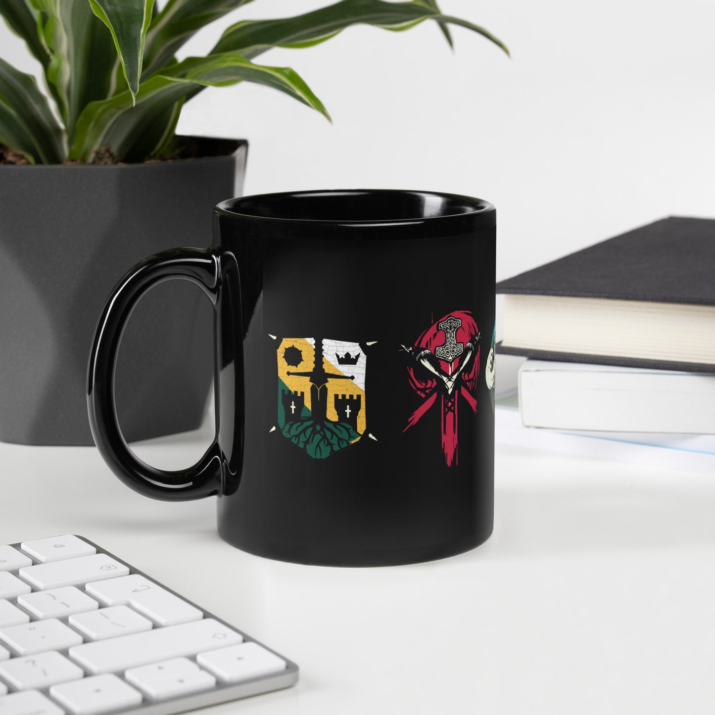 A black mug with colorful medieval-style and Viking-themed symbols sits on a white desk next to a plant, books, and a keyboar