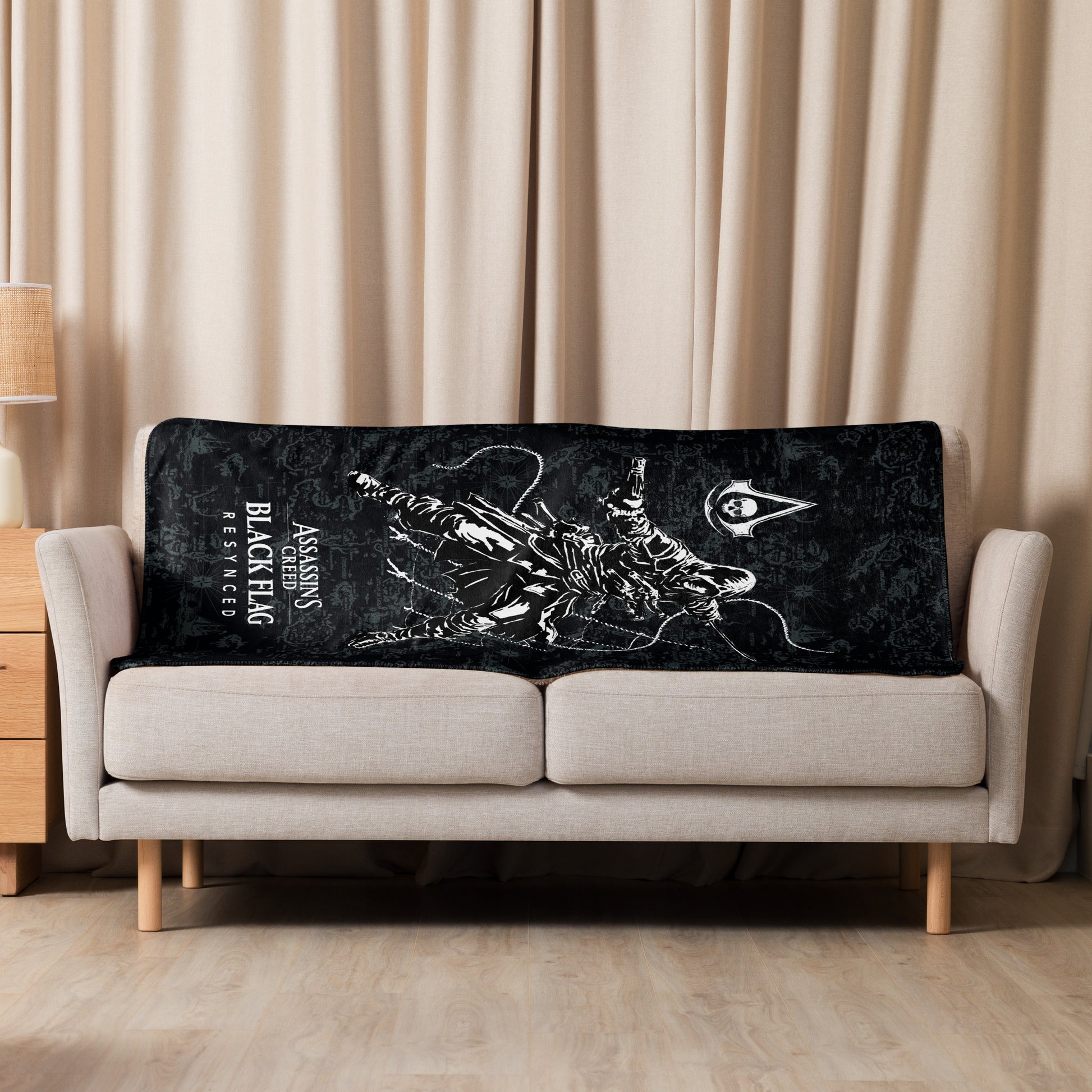 A black blanket with a white Assassin's Creed Black Flag graphic is draped over a beige two-seater sofa in a room with light 