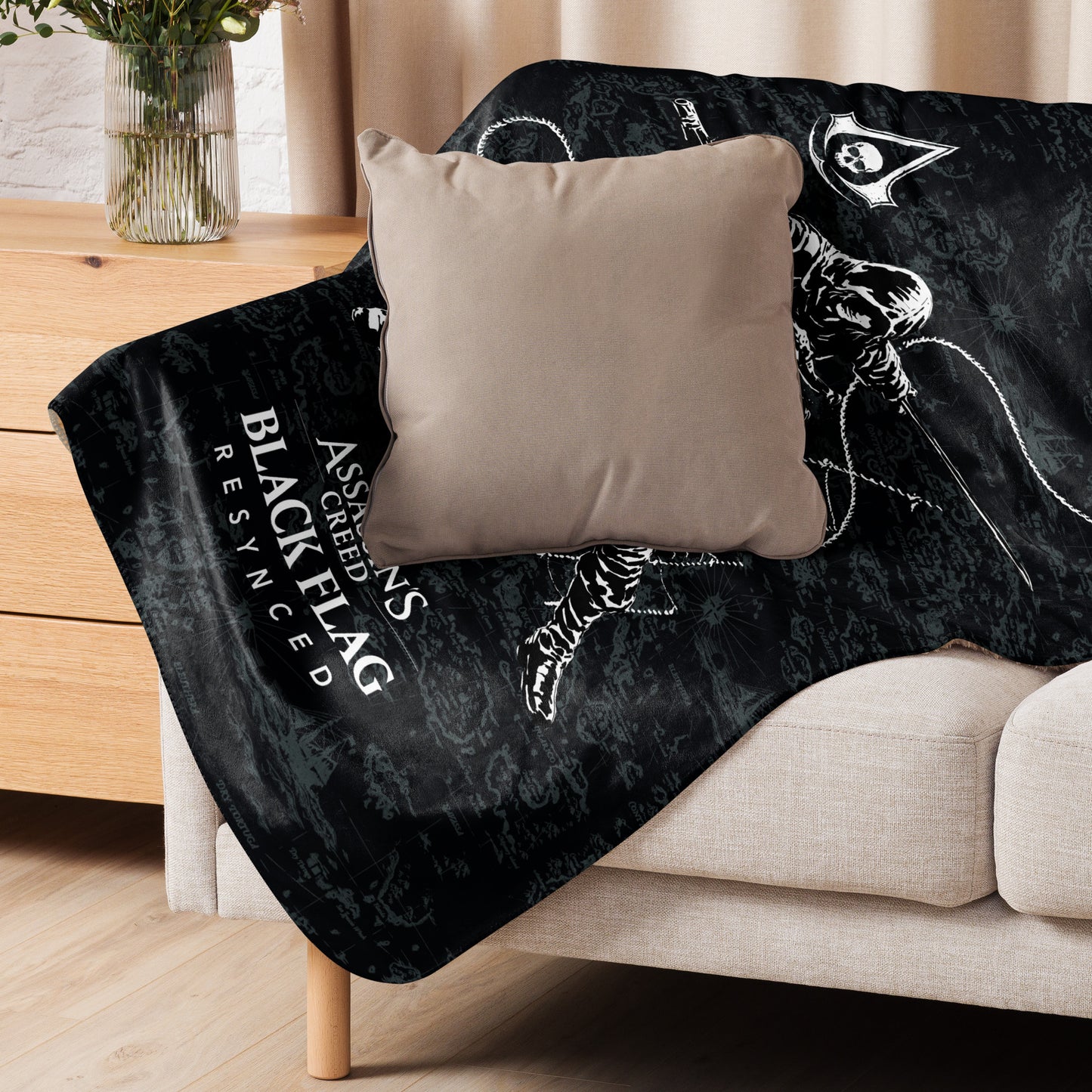 Black blanket with "ASSASSIN'S CREED BLACK FLAG RESYNCED" text and pirate map on couch, with tan pillow. Vase on table.