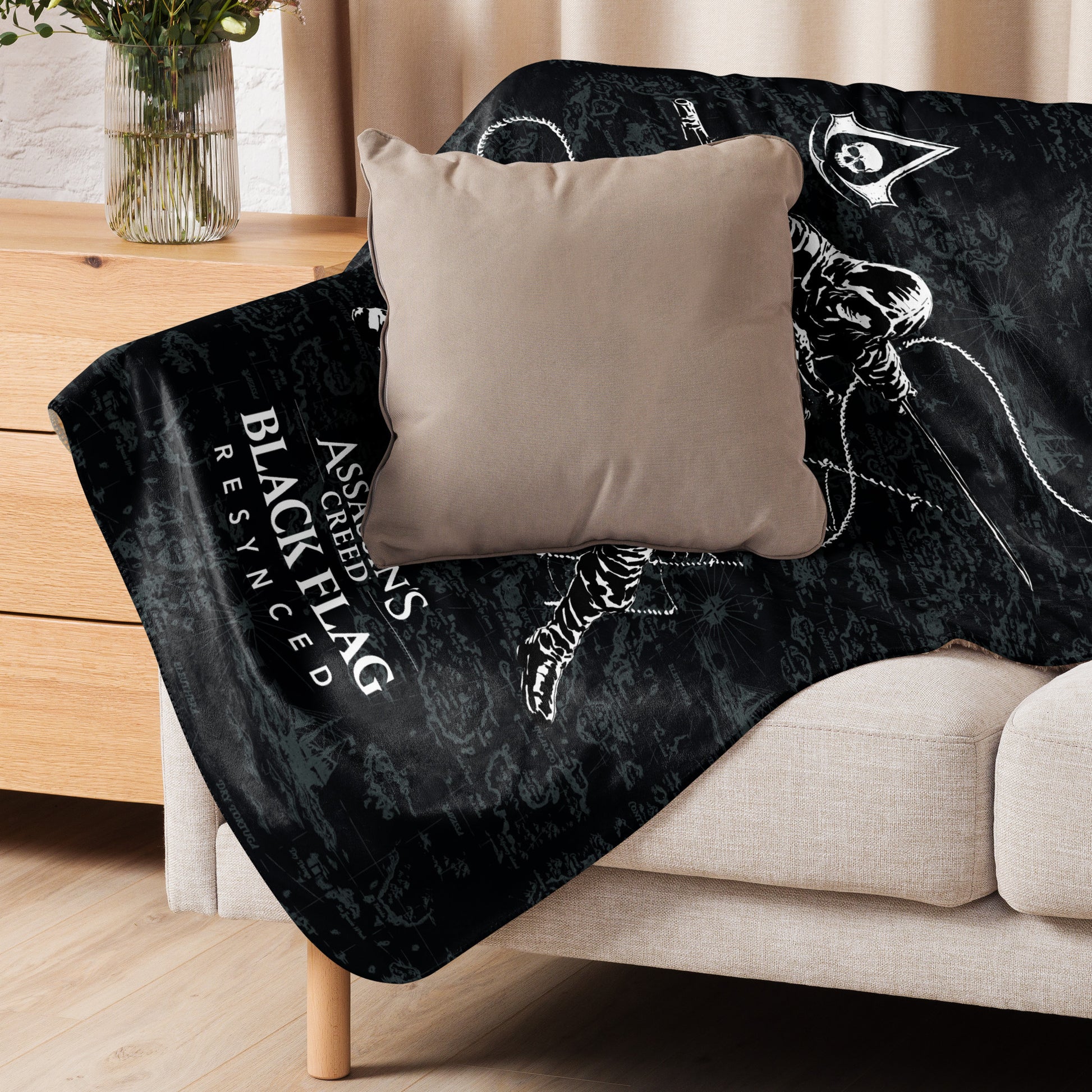 Black blanket with "ASSASSIN'S CREED BLACK FLAG RESYNCED" text and pirate map on couch, with tan pillow. Vase on table.